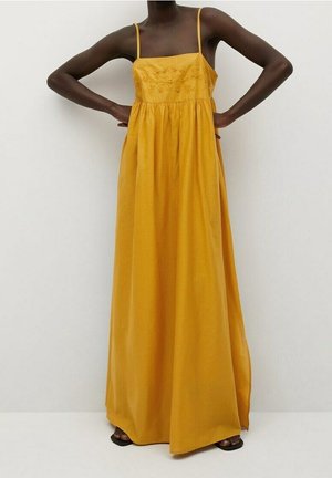 Personne portant une robe longue sans manches jaune moutarde avec de fines bretelles et un bustier brodé, debout les mains sur les hanches.