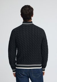 Svart kabelstickad tröja med randig krage och fåll. Har texturerat mönster och ribbade muddar. Bärs över blå jeans.