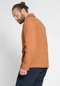 Tansfarbene Lederjacke mit Kragen, sichtbaren Nähten, seitlichen Reißverschlüssen und lässiger Passform; getragen über dunklen Hosen.