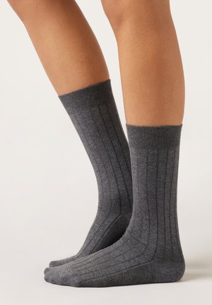 Person trägt graue, wadenhohe Rippensocken und steht mit flachen Füßen auf dem Boden vor einem schlichten, hellen Hintergrund.