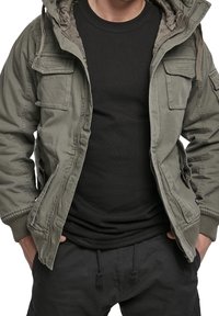 Oliwkowo-zielona kurtka w stylu wojskowym z kapturem, wieloma naszywanymi kieszeniami, zamkiem błyskawicznym i ściągaczami przy rękawach, noszona na czarnej koszulce i ciemnych spodniach.