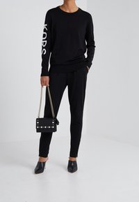 Zwarte trui met witte "KORS" letters op de mouw, gecombineerd met zwarte broek. Een kleine zwarte handtas met zilveren studs en een kettingriem.