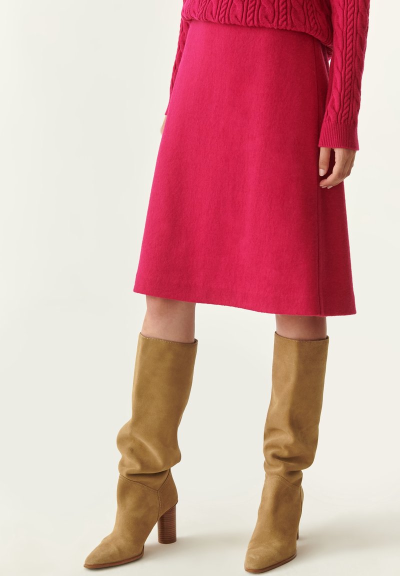 Femme portant une jupe rose vif au genou, un pull rouge à torsades et des bottines camel en daim à talons souples.
