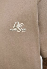 Beige bomulds t-shirt med en blød tekstur, der har et hvidt broderet logo, som står "OK originals Studio 1990" på fronten.