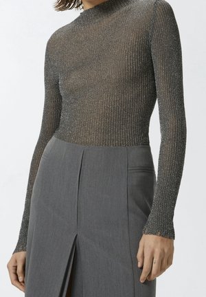 Femme portant un haut métallisé côtelé ajusté à manches longues, rentré dans un pantalon gris taille haute, coupe ajustée, avec une fente à l'avant.
