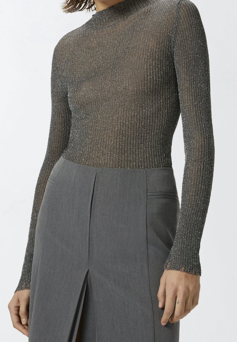 Femme portant un haut métallisé côtelé ajusté à manches longues, rentré dans un pantalon gris taille haute, coupe ajustée, avec une fente à l'avant.