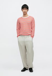 Κόκκινο και λευκό ριγέ μακρυμάνικο πουκάμισο με στρογγυλή λαιμόκοψη, σε συνδυασμό με ανοιχτό γκρι λινά παντελόνια και μαύρα παπούτσια. Μινιμαλιστικός σχεδιασμός.