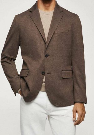 Mand iført en brun to-knaps blazer over en beige sweater med hvide bukser, den ene hånd i lommen og den anden ned langs siden.