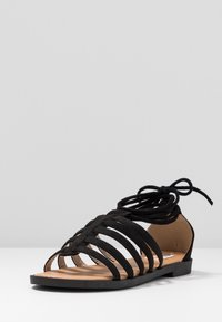 Sandale noire à lanières avec des straps en tissu tressé, une semelle plate et un design à lacets. La semelle intérieure beige complète l'extérieur noir.