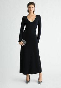 Schwarzes, langärmeliges Kleid mit tiefem V-Ausschnitt, tailliertem Oberteil und fließendem Rock. Ausgestattet mit silbernem Besatz entlang des Ausschnitts und einer strukturierten schwarzen Clutch.