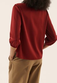 Rote Satinbluse mit drei Viertel Puffärmeln, gerafften Manschetten und abgerundetem Saum, kombiniert mit lockeren, khakifarbenen Hosen. Minimalistisches Design.