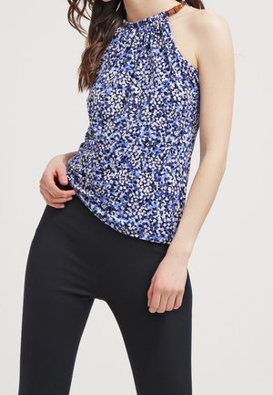 Vrouw draagt een mouwloos blauw bloemen haltertopje in combinatie met nauwsluitende zwarte broek tegen een effen lichte achtergrond.