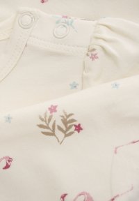 Krämfärgad tyg med blommönster i rosa och ljusblått. Inkluderar tryckknappar vid axeln för enkel påtagning. Mjuk textur.