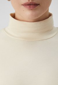 Světlý béžový rolák vyrobený z měkké, elastické látky; přiléhavý design se hladkou texturou a vysokým límcem.