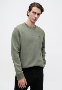 Mlad moški s kratkimi valovitimi lasmi, oblečen v olivno zeleni pulover z dolgimi rokavi in črnimi hlačami, stoji z eno roko v žepu.