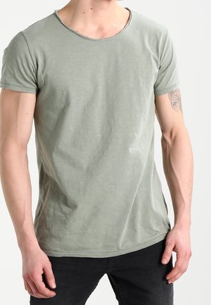 Mężczyzna w jasnozielonej koszulce z krótkim rękawem i czarnych jeansach, widoczny tatuaż na górnej części prawego ramienia, stoi na jednolitym tle.