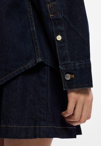 Σκούρο τζιν μπουφάν στη γωνία με ορατές ραφές, ένα μικρό κουμπί κ αι μια μεταλλική λεπτομέρεια στον καρπό; λεία υφή, κομψή γραμμή.