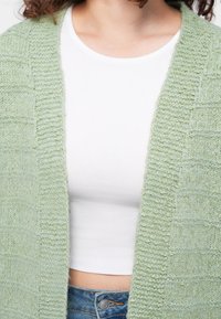 Personne portant un haut court blanc sous un cardigan ouvert texturé vert clair et un jean en denim bleu.