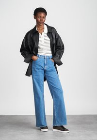 Μαύρο δερμάτινο oversize τζάκετ, λευκή και μαύρη ριγέ πόλο μπλούζα, ανοιχτό μπλε παντελόνι καμπάνα και σκούρες μονοχρωμίες παπούτσια σε απλό φόντο.