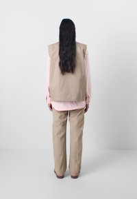 Beige ermeløs vest over en rosa langermet skjorte, parret med lyse beige, løstsittende bukser. Glatt tekstur, enkel design.