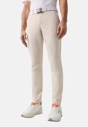 Mand iført beige, tætsiddende bukser, hvidt bælte og hvide sneakers med orange snørrebånd, stående mod en ensfarvet baggrund.