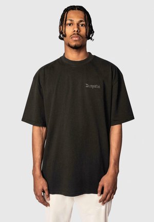 Svart oversized t-skjorte laget av bomull, med korte ermer og en liten, brodert logo på brystet. Enkelt, rent design.