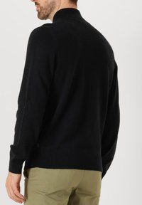 Pull en maille noire avec un col haut, texture douce et coupe décontractée. Associé à un pantalon vert clair pour un look décontracté.