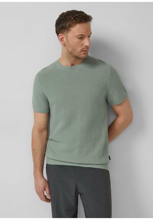 Mann trägt einen kurzärmligen, strickgrünen Pullover in Salbeigrün und dunkelgraue Hosen, blickt nach unten und zur Seite vor einem einfarbigen Hintergrund.