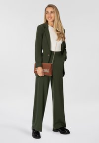 Olijfgroene op maat gemaakte pak met een ontspannen pasvorm, voorzien van een blazer met één knoop en brede broekspijpen; gecombineerd met een bruine schoudertas.