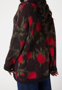 Maglione oversize in lana scura, con un motivo floreale rosso e accenti verdi, polsini e orlo a coste, texture morbida e vestibilità rilassata.