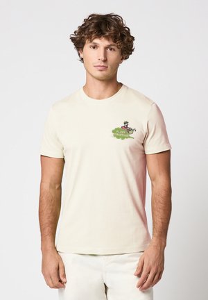 Jeune homme aux cheveux bouclés portant un t-shirt écru avec un petit motif vert "Plant Together" et un arrosoir sur la poitrine.