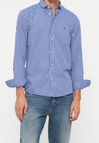 Homme portant une chemise à manches longues à carreaux bleu et blanc avec les manches retroussées et un jean denim bleu clair, debout devant un fond uni.