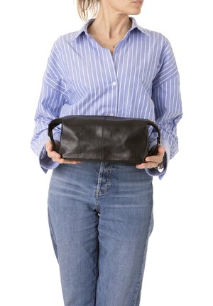 Femme portant une chemise à rayures bleues et un jean, tenant devant elle une grande pochette en cuir noire avec des poignées.