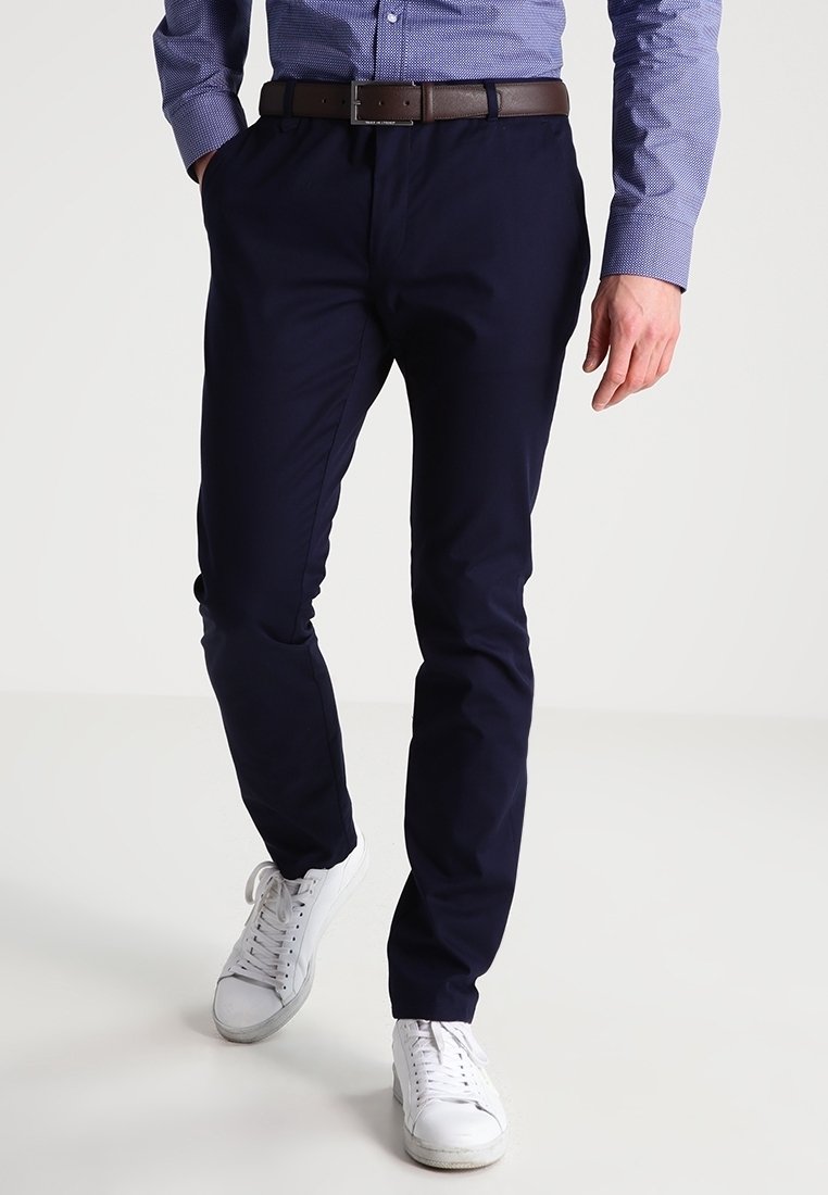 Homme portant un pantalon bleu marine foncé, une ceinture marron, une chemise bleue à motifs et des baskets blanches, marchant devant un fond uni.