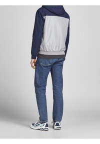 Navy and gray hooded jacket with elastic hem, worn over a dark layer, paired with blue denim jeans and white sneakers with navy accents.