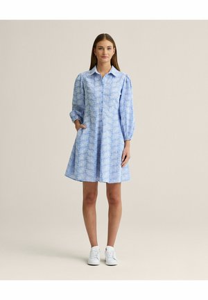 Kvinde stående iført en lyseblå blomsterprintet langærmet kjole og hvide sneakers, med den ene hånd i lommen mod en ensfarvet beige baggrund.