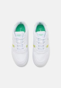 Sneaker bianche con tomaia sintetica liscia, dotate di una striscia gialla, soletta verde e suola testurizzata per la presa. Etichettate con "Lacoste."