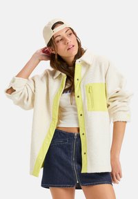 Cremefarbene Fleecejacke mit einer leuchtend gelben Druckknopfleiste und Tasche. Strukturierter Stoff und hochgerollte Ärmel. Getragen mit einem Denim-Rock.