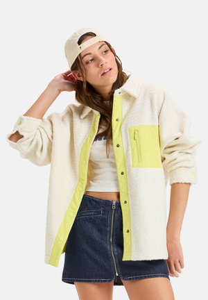 Veste en polaire crème avec une patte de boutonnage à pression jaune vif et une poche. Tissu texturé et manches retroussées. Portée avec une jupe en jean.