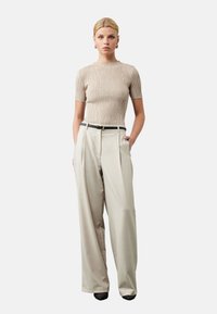 Blonde vrouw in een beige textuur kortemouwen top, hooggewaardeerde beige broek met wijde pijpen en een zwarte riem, staand met haar handen in de zakken.
