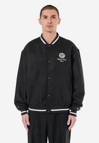 Sort varsityjakke med hvidstribede krave og manchetter, knaplukning og broderet detalje på brystet. Blødt, tekstureret stof.