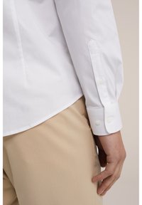 White button-up shirt with a smooth texture, featuring a rounded collar, buttoned cuffs, and a straight hem, paired with beige trousers.