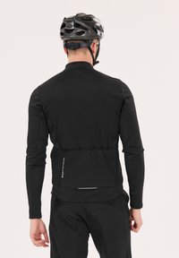 Veste de cyclisme noire à manches longues avec une finition texturée, des accents réfléchissants et une poche arrière. Portée avec un casque de vélo noir.