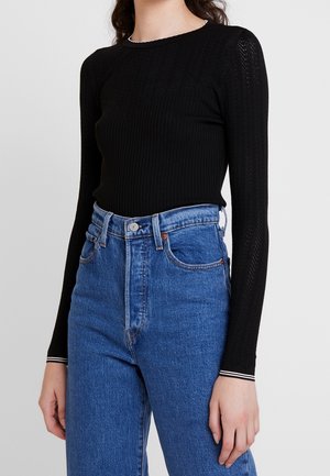 Vrouw draagt een nauwsluitend zwart shirt met lange mouwen en een textuurpatroon, ingestopt in een hoogwater-blauwe spijkerbroek, tegen een effen achtergrond.
