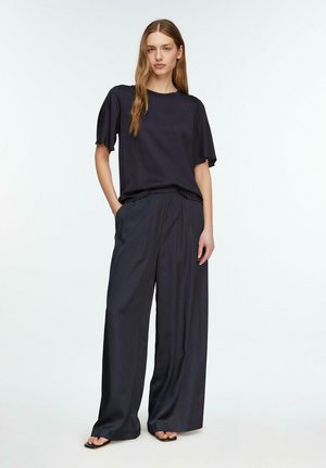 Kobieta o długich włosach stoi, ubrana w luźną granatową bluzkę z krótkim rękawem i szerokie spodnie oraz sandały, na tle jednolitego jasnego tła.