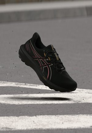 Chaussure de running ASICS noire avec des touches roses en plein air au-dessus d’une surface en asphalte gris avec des lignes blanches peintes.