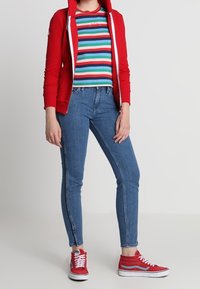 Rode hoodie met rits over een meerkleurig gestreept t-shirt, gecombineerd met blauwe skinny jeans en rode hoge sneakers.