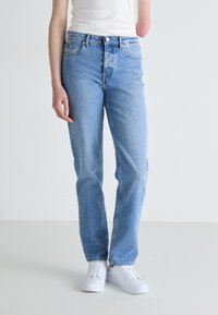 Light blue denim jeans with a straight-leg design, five-pocket styling, and a button-and-zip closure. Paired with white sneakers.