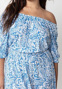 Kvinna klädd i en axelbar blå och vit blommönstrad klänning med puffärmar och elastisk midja.