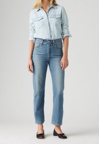 Camisa de mezclilla clara con dos bolsillos en el pecho, combinada con jeans azules de tiro alto. Bailarinas negras con detalle de lazo complementan el atuendo.